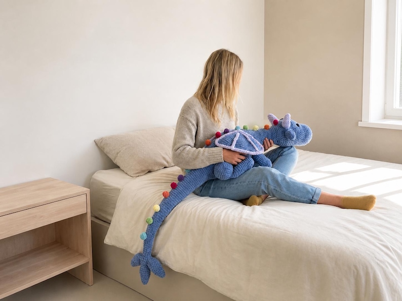 May include: A large, blue crocheted dragon plush toy with rainbow pom-pom accents along its tail and back. The dragon has a detailed head and wings. The toy is soft and cuddly, perfect for children or as a decorative item.