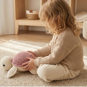May include: A handmade crochet turtle plush toy with a white head and limbs, and a mauve shell. The turtle is on a beige rug. The image shows a child holding the turtle. The toy is a soft, cuddly item, perfect for a nursery or child's room.