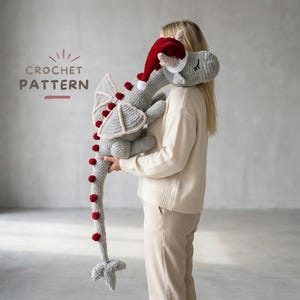 May include: A crocheted gray dragon toy with red accents and a Santa hat. The dragon has large wings and a long tail with red pom-poms. The text "CROCHET PATTERN" is visible in the upper left corner.