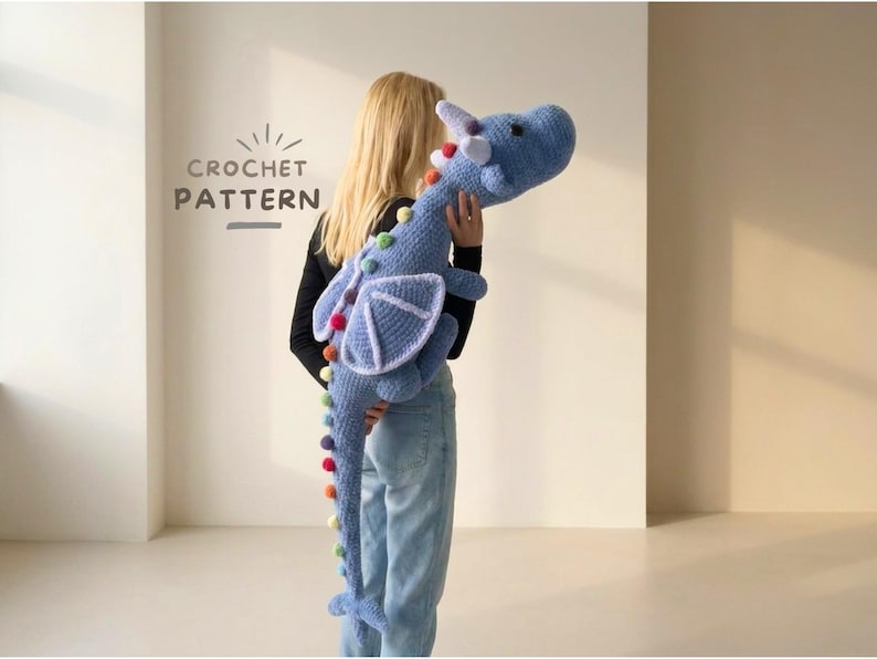 May include: A large, light blue crochet dragon with a white horn, wings, and colorful pom-poms along its back and tail. The dragon is held by a person. The text "CROCHET PATTERN" is visible in the upper left corner.