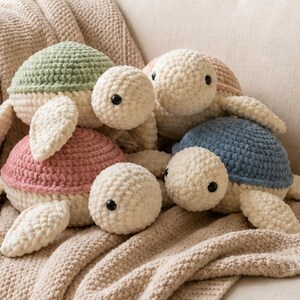 May include: A collection of handmade crochet turtle plushies in various pastel colors. The turtles have cream-colored bodies, flippers, and heads, with shells in shades of green, pink, blue, and peach. They are arranged on a beige knitted blanket.
