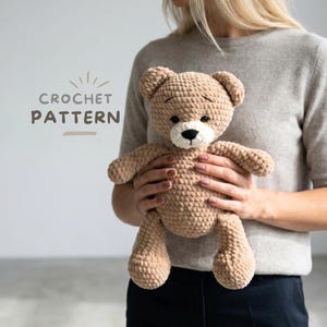 May include: A crocheted teddy bear in a light brown color, with a white muzzle and black eyes. The bear is held by a person. The text "CROCHET PATTERN" is in the upper left corner. The bear is a handmade item.