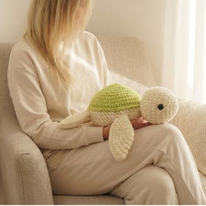 May include: A handmade crochet turtle plush toy. The turtle has a light green shell, cream-colored body, and a black button eye. The turtle is a soft, cuddly toy, perfect for children or as a decorative item. The turtle is approximately 12 inches long.