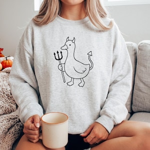 May include: A light gray crewneck sweatshirt featuring a black line art graphic of a duck with small horns, a pointed tail, and a trident. The duck is depicted in a simple, cartoon-like style centered on the chest of the long-sleeved garment.