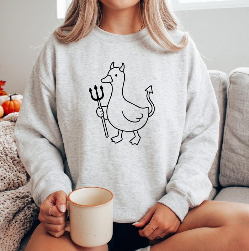 May include: A light gray crewneck sweatshirt featuring a black line art graphic of a duck with small horns, a pointed tail, and a trident. The duck is depicted in a simple, cartoon-like style centered on the chest of the long-sleeved garment.