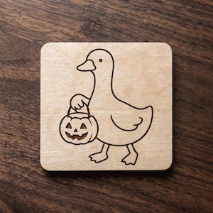 May include: A square light-colored wooden coaster featuring a dark laser-engraved line art illustration of a duck holding a carved jack-o'-lantern pumpkin bucket. The design is centered on the smooth surface of the natural wood material.