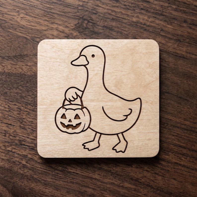 May include: A square light-colored wooden coaster featuring a dark laser-engraved line art illustration of a duck holding a carved jack-o'-lantern pumpkin bucket. The design is centered on the smooth surface of the natural wood material.
