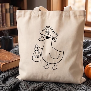 May include: A cream-colored canvas tote bag featuring a black line art illustration of a duck dressed as a pirate. The duck wears a pirate hat with a skull and crossbones symbol and an eye patch, while holding a small sack of candy in its wing.