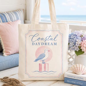 May include: A cream-colored tote bag with a coastal theme. The bag features a graphic of a seagull perched on a pink and white striped post, with the words "Coastal Daydream" in blue script. The background includes a pink circle and blue waves.