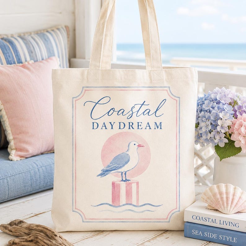 May include: A cream-colored tote bag with a coastal theme. The bag features a graphic of a seagull perched on a pink and white striped post, with the words "Coastal Daydream" in blue script. The background includes a pink circle and blue waves.
