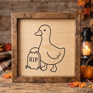 May include: A square wooden sign with a dark brown frame featuring a black line art illustration of a duck standing next to a tombstone with the text RIP engraved on the light-colored wood surface. The design is centered on the flat wooden panel.