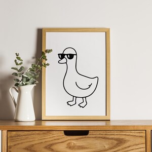 May include: A framed print featuring a minimalist line drawing of a duck wearing sunglasses. The artwork is in black and white, set against a white background, and framed in a light wood frame. The print is displayed on a wooden surface.