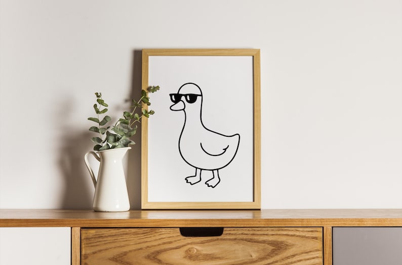 May include: A framed print featuring a minimalist line drawing of a duck wearing sunglasses. The artwork is in black and white, set against a white background, and framed in a light wood frame. The print is displayed on a wooden surface.