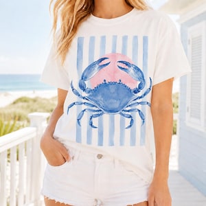 May include: White t-shirt featuring a blue crab design with a pink sun and blue vertical stripes. The crab is detailed with blue claws and legs. The shirt is paired with white shorts.