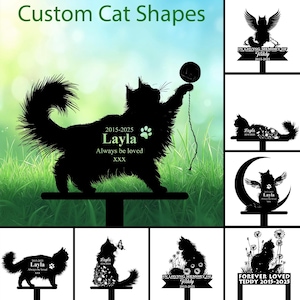 May include: Black metal cat-shaped memorial stakes. The main stake features a cat silhouette playing with a ball, with the name "Layla" and dates. Other stakes include cat silhouettes with angel wings, flowers, and the text "Forever Loved".