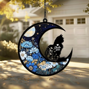 May include: A circular hanging ornament shaped like a crescent moon featuring a black silhouette of a cat, a white heart, and the text Charlie 2025. The design includes blue and white floral patterns, a dark blue starry sky, and a gold moon.
