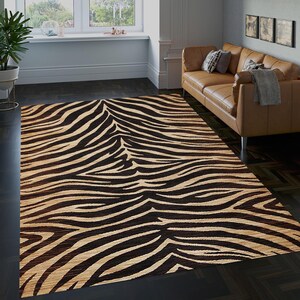 Puede incluir: Una alfombra rectangular con un diseño de rayas de cebra. La alfombra presenta bandas alternas de marrón oscuro y beige, creando un diseño audaz de estampado animal. La alfombra está colocada sobre un suelo de madera oscura en un salón.