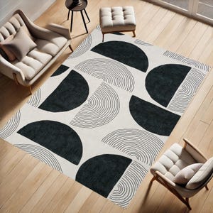 Op de afbeelding: Een groot, rechthoekig vloerkleed met een geometrisch ontwerp. Het vloerkleed heeft grote, zwarte halve cirkels en gebogen lijnen tegen een crèmekleurige achtergrond. Het vloerkleed ligt op een houten vloer en is omringd door meubels.