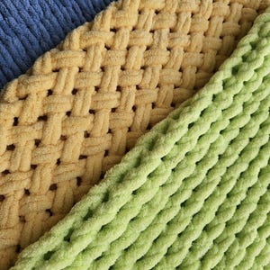 Puede incluir: Primer plano de tres mantas de punto en diferentes colores: una manta azul claro, una manta amarilla con un patrón tejido y una manta verde con un patrón de trenza.