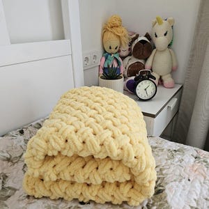 Pu&ograve; includere: Una coperta di lana gialla piegata su un letto con un copriletto a motivi floreali. La coperta &egrave; realizzata con filato spesso e ha una trama a maglia grossa.
