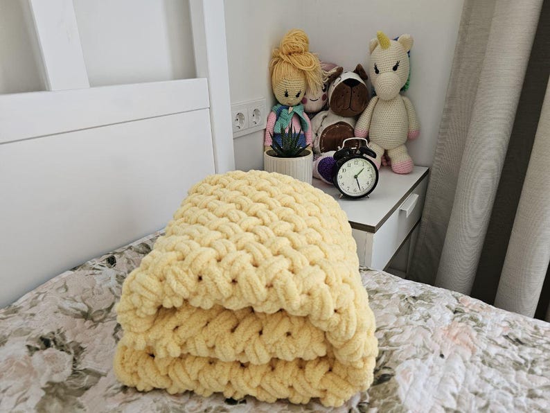 Pu&ograve; includere: Una coperta di lana gialla piegata su un letto con un copriletto a motivi floreali. La coperta &egrave; realizzata con filato spesso e ha una trama a maglia grossa.