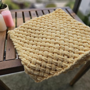 Pu&ograve; includere: Una coperta di maglia gialla con una trama tessuta. La coperta &egrave; drappeggiata su un tavolo di legno.