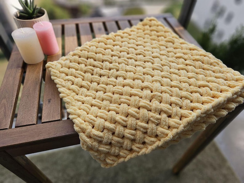 Pu&ograve; includere: Una coperta di maglia gialla con una trama tessuta. La coperta &egrave; drappeggiata su un tavolo di legno.