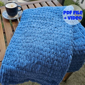 Puede incluir: Una manta de punto azul con un patrón texturizado. La manta está doblada y colocada sobre una mesa de madera.