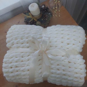 Könnte beinhalten: Eine weiße gestrickte Wolldecke mit einer großen, flauschigen Textur. Die Decke ist mit einem weißen Satinband gebunden.