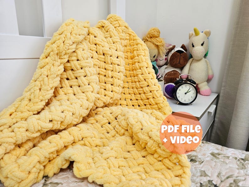 Pu&ograve; includere: Una coperta di maglia gialla con un motivo intrecciato. La coperta &egrave; su un letto con una testiera bianca e un comodino con un orologio e animali di peluche.
