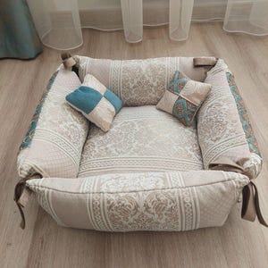May include: A beige and white patterned dog bed with a removable cushion and two decorative pillows. The pillows are also patterned with beige and white, and one has a blue and white checkered design.