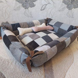 Puede incluir: Una cama para perro de patchwork gris y blanco con una almohada en forma de hueso. La cama tiene un lazo de cuero marrón en cada esquina.