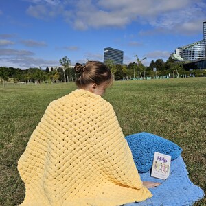 Pu&ograve; includere: Una persona &egrave; seduta su una coperta all'uncinetto blu e gialla in un parco erboso. La persona sta leggendo un libro intitolato "Help" con un cuscino blu accanto a lei.