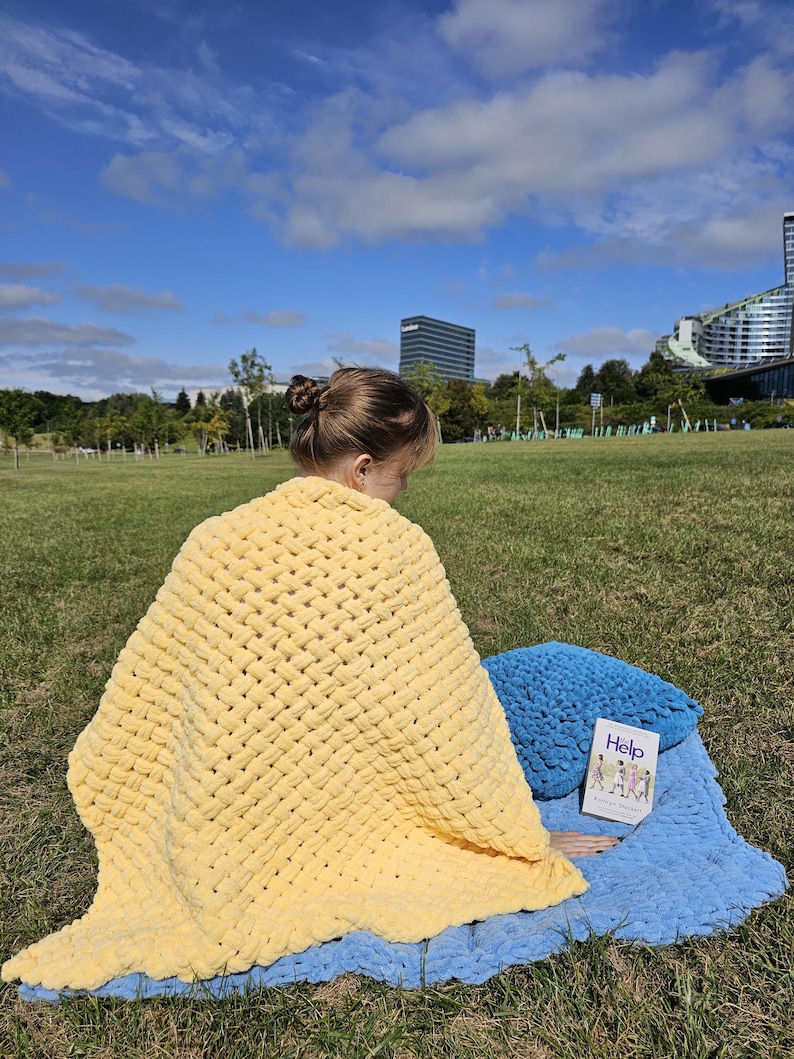 Pu&ograve; includere: Una persona &egrave; seduta su una coperta all'uncinetto blu e gialla in un parco erboso. La persona sta leggendo un libro intitolato "Help" con un cuscino blu accanto a lei.