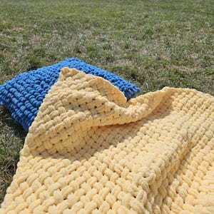 Pu&ograve; includere: Una coperta di maglia gialla e blu con una trama a maglia grossa. La coperta &egrave; piegata e drappeggiata su un cuscino di maglia blu.