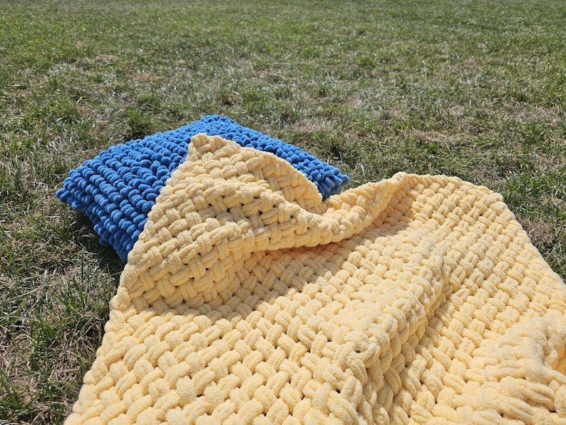 Pu&ograve; includere: Una coperta di maglia gialla e blu con una trama a maglia grossa. La coperta &egrave; piegata e drappeggiata su un cuscino di maglia blu.