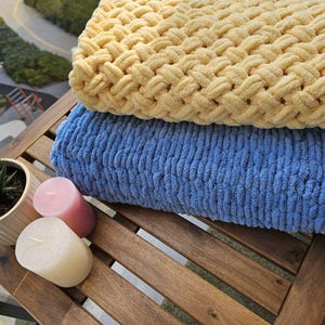 Pu&ograve; includere: Due coperte di maglia, una gialla e una blu, sono piegate su un tavolo di legno con due candele.