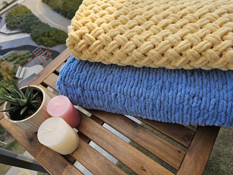Pu&ograve; includere: Due coperte di maglia, una gialla e una blu, sono piegate su un tavolo di legno con due candele.