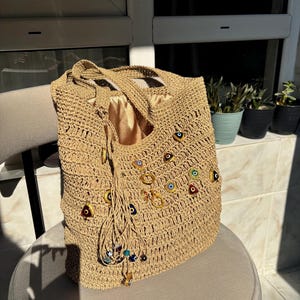 May include: A beige crocheted tote bag featuring a satin lining and multiple small, colorful evil eye charms attached to the front. The bag includes two shoulder straps and a decorative tassel made of matching yarn with additional small beads.
