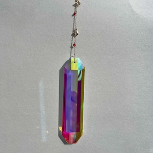 Op de afbeelding: Een prismavormige kristallen hanger met een gouden ketting en steraccenten. Het kristal reflecteert een spectrum aan kleuren, waaronder roze, paars en geel. Kleine rode kralen zijn langs de ketting verspreid. De hanger hangt tegen een witte achtergrond.