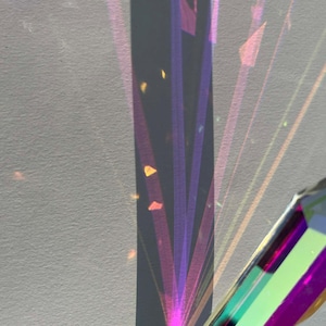 Op de afbeelding: Een close-up van een kristallen prisma dat licht breekt en een spectrum van kleuren creëert. Het licht wordt op een wit oppervlak geprojecteerd en vormt levendige lijnen en vormen. Het prisma reflecteert tinten paars, groen en oranje.