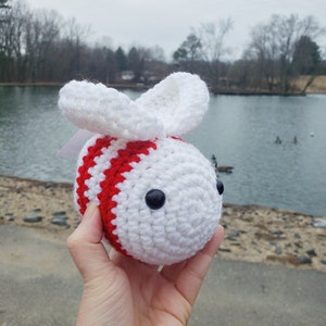 May include: A white crocheted bee with red stripes and black eyes. The bee is being held in a hand.