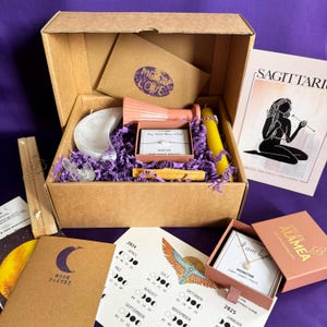 Könnte beinhalten: Eine braune Pappbox gefüllt mit verschiedenen Artikeln, darunter eine Mondstein-Halskette, ein mondförmiger Kristall, eine Kerze, eine kleine rosa Vase, ein Holzstab und eine Karte mit einem Mondkalender. Die Box ist geöffnet und die Artikel sind sichtbar. Die Box ist mit "Moon Love" beschriftet.