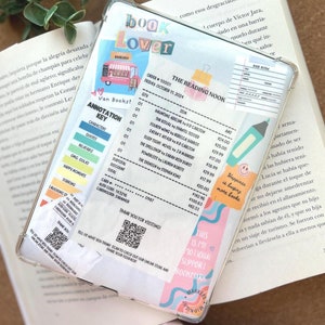 May include: A clear phone case with a pink and blue book lover theme. The case has a sticker of a book shop with a red truck and the words "Book Lover" in colorful letters. The case also has a sticker with a list of books and their prices. The case has a pink and blue bookmark with the words "This is my motivational support bookmark".