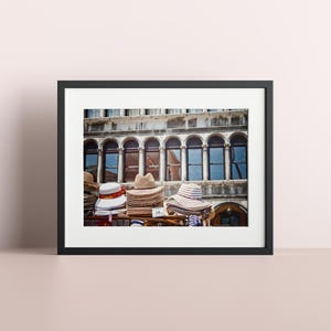 May include: A framed photograph of a stack of straw hats in front of a building with arched windows. The building is made of stone and has a white facade. The hats are various colours and styles.