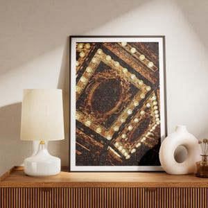 May include: A framed art print featuring a detailed, abstract design in shades of brown and gold. The artwork has a vintage aesthetic, with ornate, repeating patterns. A white lamp and vase sit on a wooden surface.
