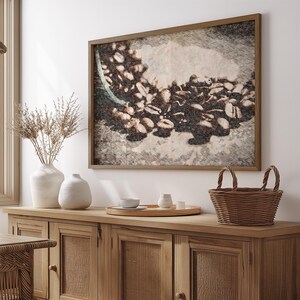 Puede incluir: Una impresión artística enmarcada que representa granos de café derramándose de un recipiente. La obra de arte está en un estilo de boceto, con tonos marrones y beige. La impresión cuelga sobre un aparador de madera con una cesta tejida y jarrones decorativos.