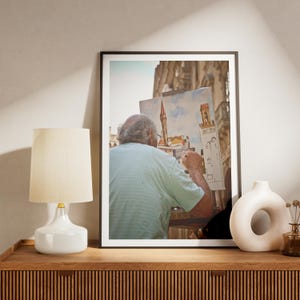 May include: A person paints a cityscape on a canvas while standing in front of a building. The painting depicts a tall tower with a pointed roof and a smaller building next to it. The person is wearing a light blue striped shirt and is holding a paintbrush in their right hand.