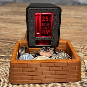 May include: A black coin-operated arcade-style box with a red illuminated display reading 25¢ INSERT COIN TO PLAY, positioned behind a brown, brick-textured plastic tray filled with several metallic silver tokens.