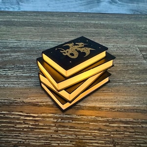 May include: A stack of three black, illuminated book-shaped lights. The top book features a gold dragon design. The pages of each book glow with a warm, yellow light. The books are stacked on a wooden surface.